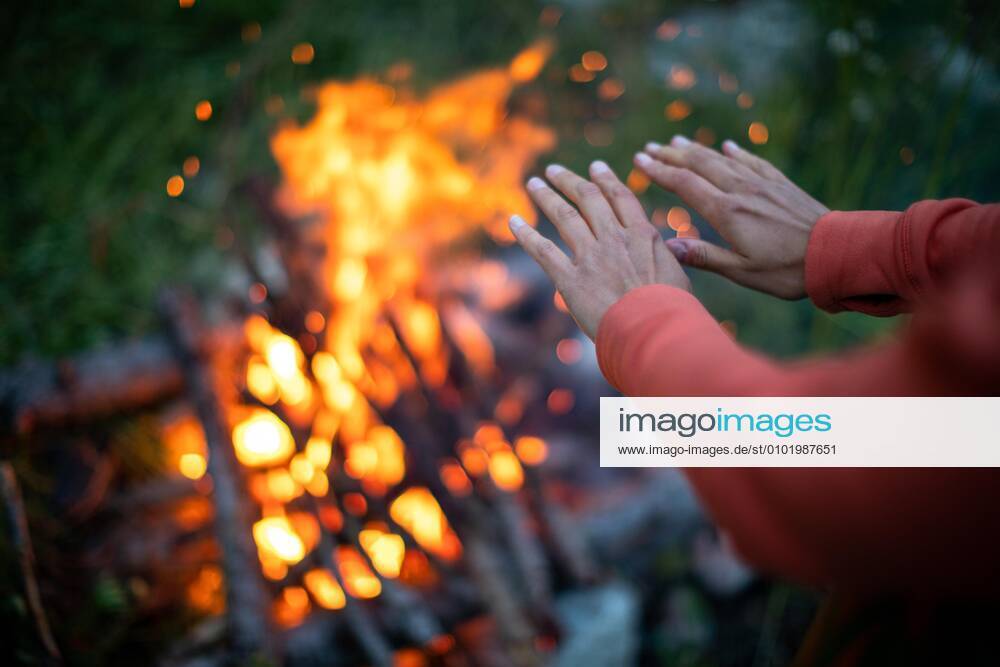 Young woman making fire while camping outdoors, in an alpine wilderness - warming up her hands near the fire