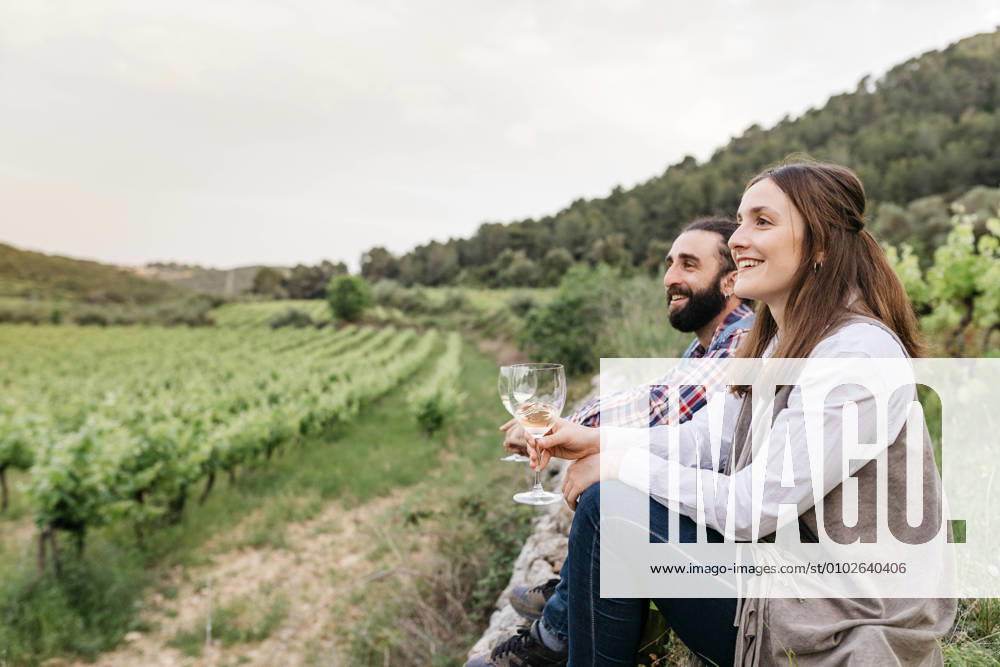 IMAGO Creative, Sommer, Weinfeste und Weinwanderungen Happy couple holding white wine glasses while sitting at vineyard model released Symbolfoto JRFF04495