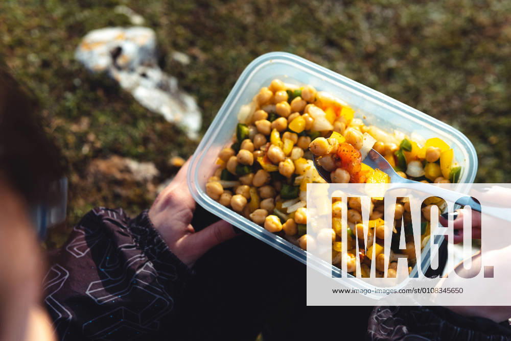 From above crop woman in warm jacket holding plastic container of chickpea with veggies having lunch on meadow while hiking in Spain, Model released