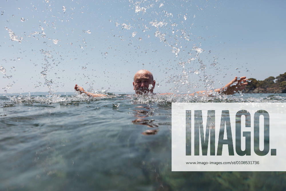 IMAGO Creative: Sommer Sport - Strand Bald mustache elderly man enjoying and splashing water looking at camera in bright sunny day in Greece Halkidiki