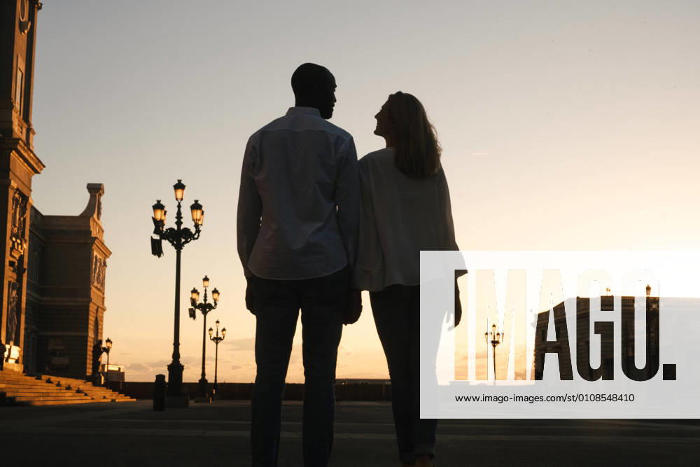 IMAGO Creative: Sommer, Abendspaziergang Back view anonymous diverse couple in casual outfits walking together along asphalt sidewalk on street during sunset time, Model released