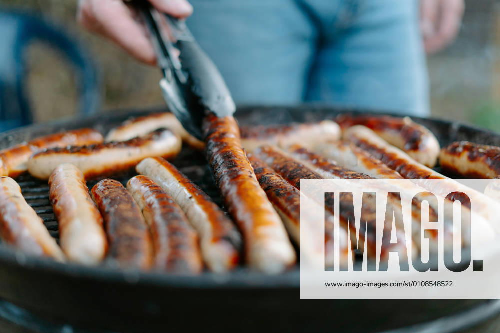 Crop anonymous person in jeans frying delicious beef sausages on grill and using tongs while having barbecue picnic on backyard