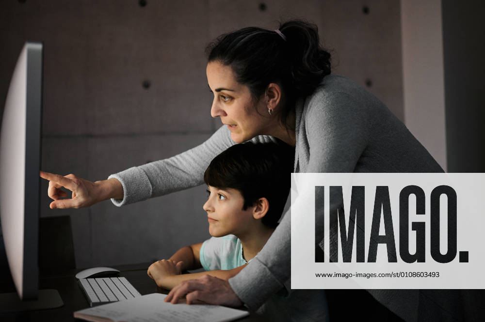 IMAGO Creative: Gesellschaft, Integration Side view of young woman explaining study task to positive son sitting at table with computer and textbook during online lesson at home, Model released