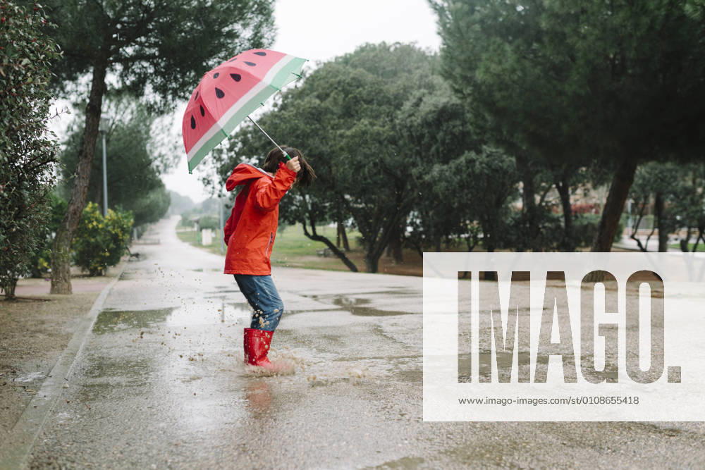 Side view active kid with watermelon styles open umbrella in red raincoat and rubber boots jumping playing in park alley in gray day, Model released