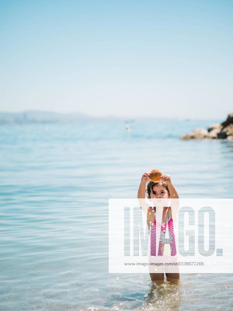 IMAGO Creative, Jahreszeit Sommer, Sommerferien Adorable toddler girl in swimming suit standing in warm water of calm sea playing with a seashell