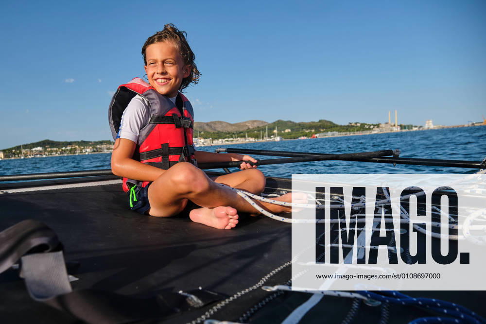Blond kid with life jacket sitting in a boat holding the rudder and a rope while smiling and the shore far away in the background, Model released