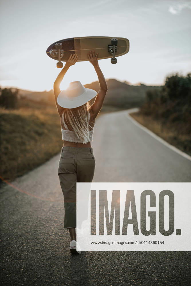 IMAGO Creative: Sommer, Abendspaziergang Back view young female in casual wear and summer hat holding cruiser skateboard and looking away while standing on empty asphalt road in rural area at sunset, Model released