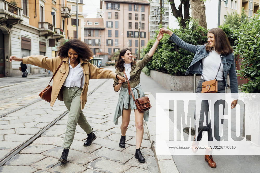 IMAGO Creative: Gesellschaft, Integration Cheerful women holding hands while walking at city street model released Symbolfoto MEUF03496