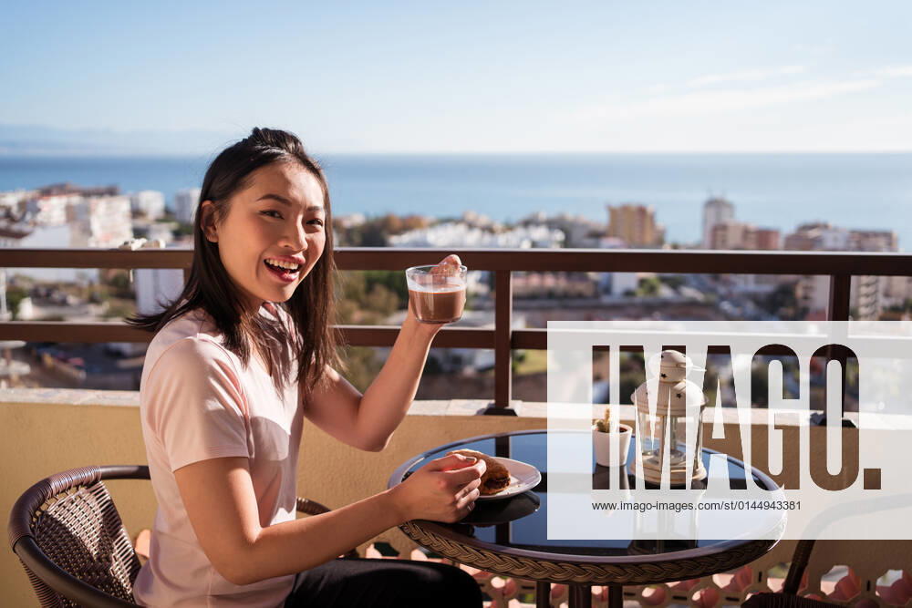 Happy Asian female with glass of hot drink in hand looking at camera while sitting on balcony at table with food during breakfast, Model released JuanAlbertoRuiz_BreakfastAtHome_7.jpg