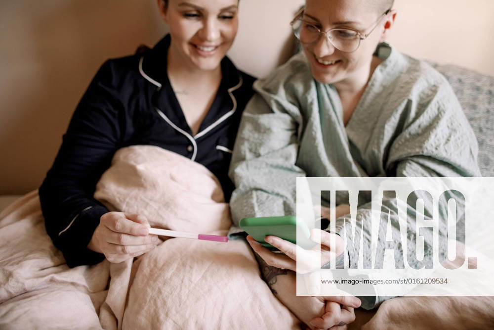 IMAGO Creative: Business, Innovative Technologie-Lösungen Happy lesbian couple checking pregnancy test results with smart phone while sitting on bed at home Sweden, Stockholm, model released, property released