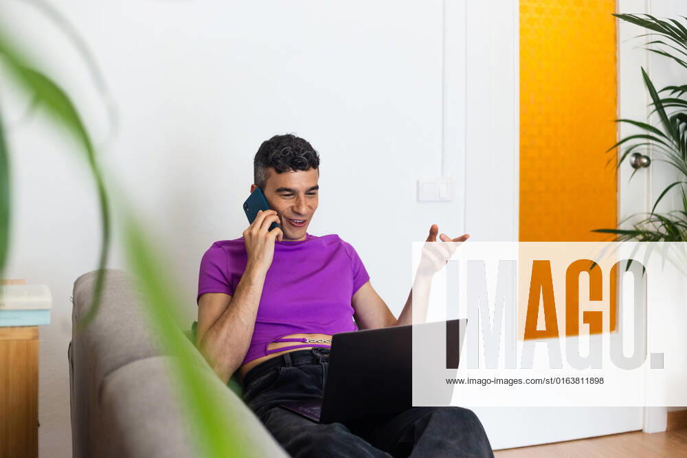 IMAGO Creative: Business, Innovative Technologie-L?sungen Smiling transgender man using phone and laptop sitting on sofa at home, Model released JordiBataller_QueerReality_48.jpg