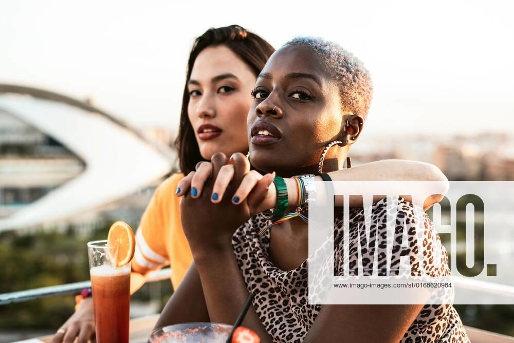 Stylish multiracial girlfriends sitting in open air cafe hugging and holding hands while enjoying refreshing drinks on blurred background, Model released RafaCortes_LesbianEthnicCouple_117