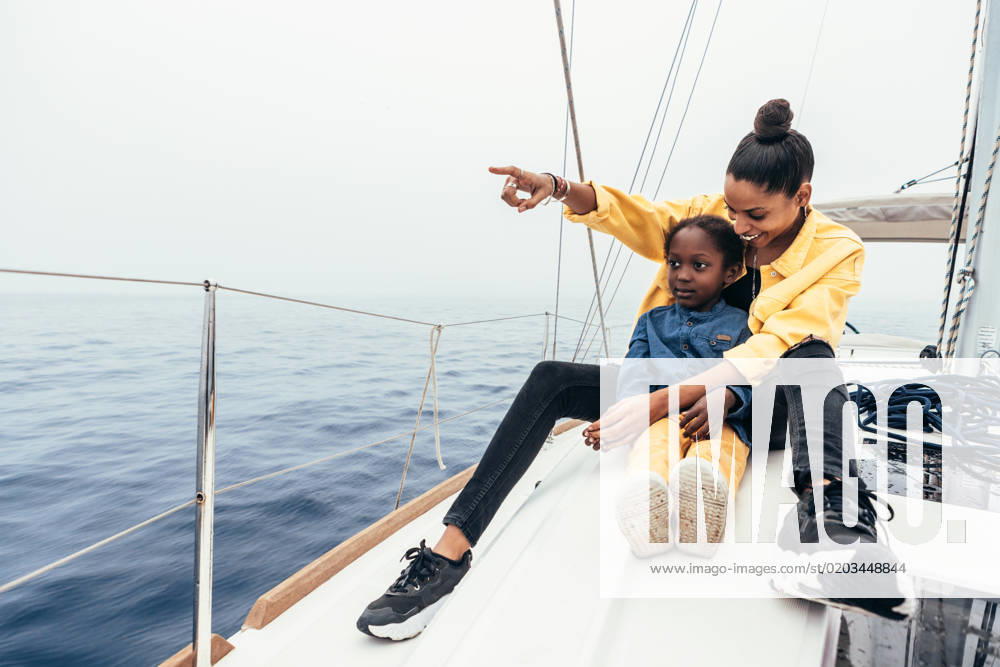 Full body of happy African American mother in casual clothes sitting on yacht and pointing away while smiling and showing view to little son during weekend, Model released AlfonsoSaenz_ID12778_387425_012