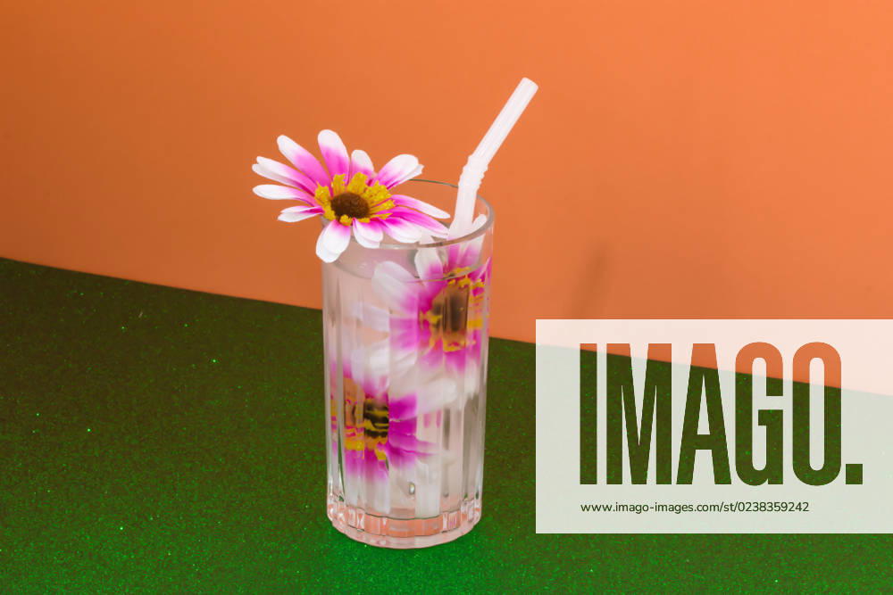 Transparent glass of refreshing cold drink decorated with pink flowers and straw placed on green surface against bright orange wall Rodobot_ID12962_394298_002