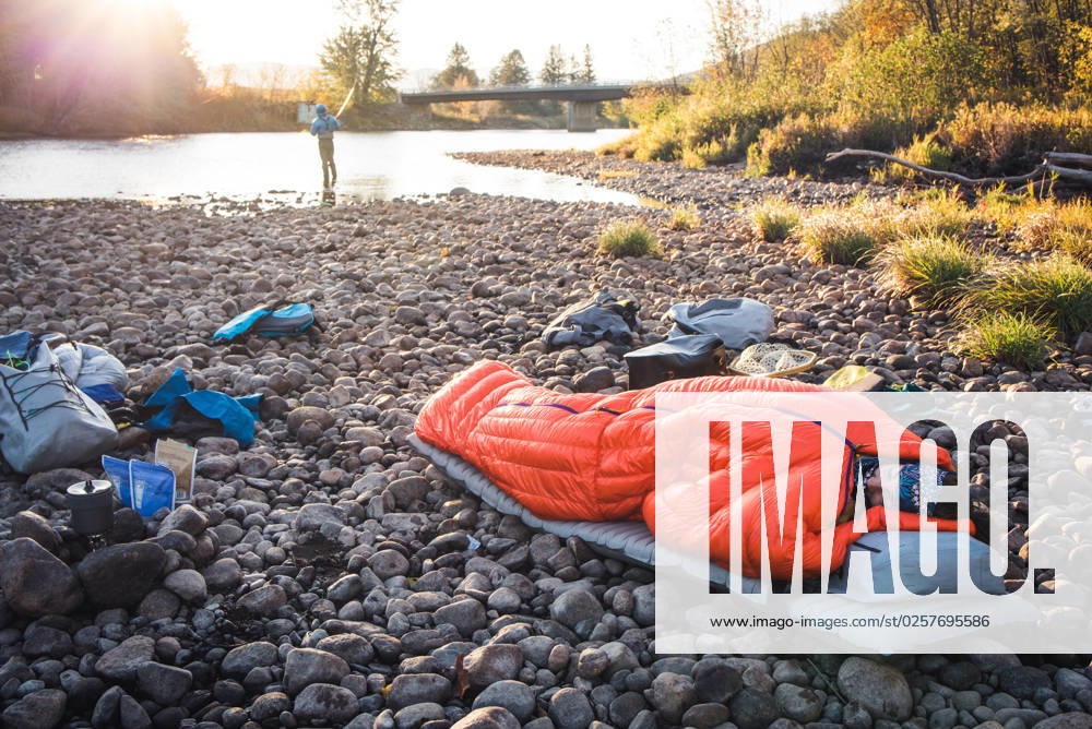 Woman asleep in sleeping bag on river edge with fiisherman behind Shelburne, New Hampshire, United States CR_BRDO2302087A-1123611-01 ,model released, Symbolfoto