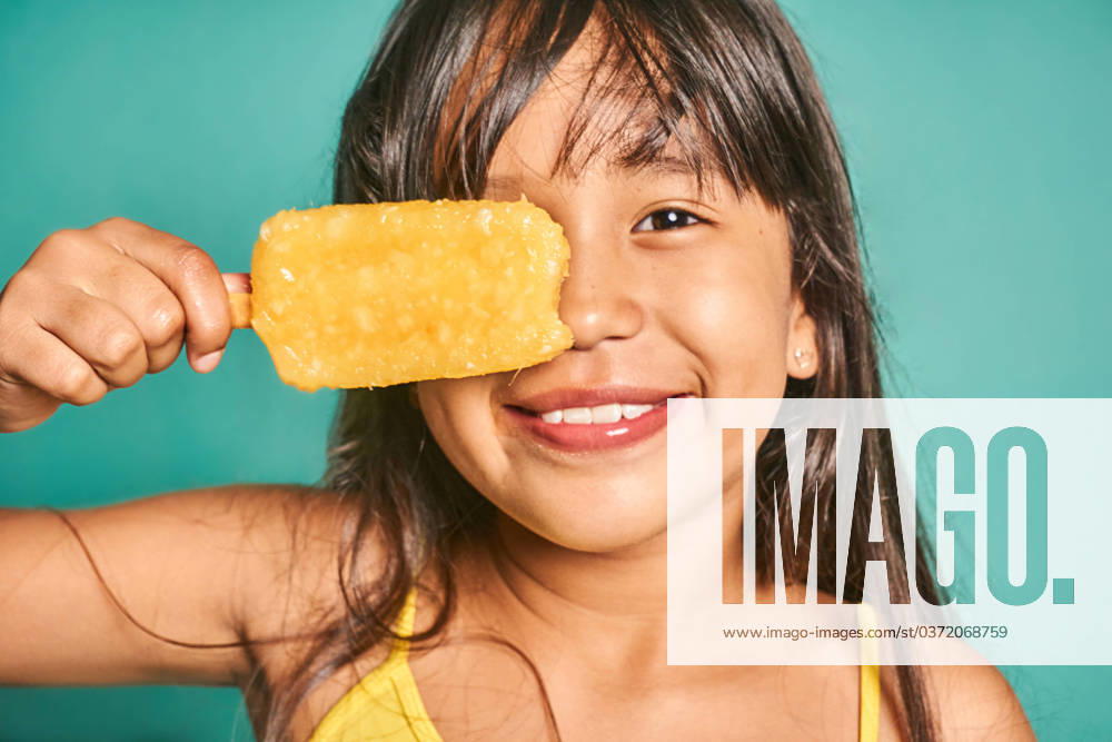 Happy ethnic girl child standing with popsicle near face against turquoise backdrop Cute crop Asian kid girl with long hair looking at camera while standing and covering eye with delicious yellow ice cream against turquoise background Model Released JoseDeJesusSaldana_ID16142_491319_034