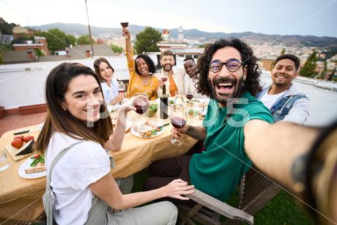 Group of multiracial friends having fun at rooftop party. Man taking selfie to people at barbecue table. Smiling companions enjoy eating and drinking on outdoor terrace. Relationships and celebrations {rooftop selfie friends happy barbecue group multiracial together party outdoors drinking diverse family people meeting food tasting eating cheerful youth alcohol barbeque bbq beverage birthday brunch celebration hangout home lifestyle lunch picnic vacation having fun taking dinner friendship time smiling terrace positive relations millennial anniversary man women beard}