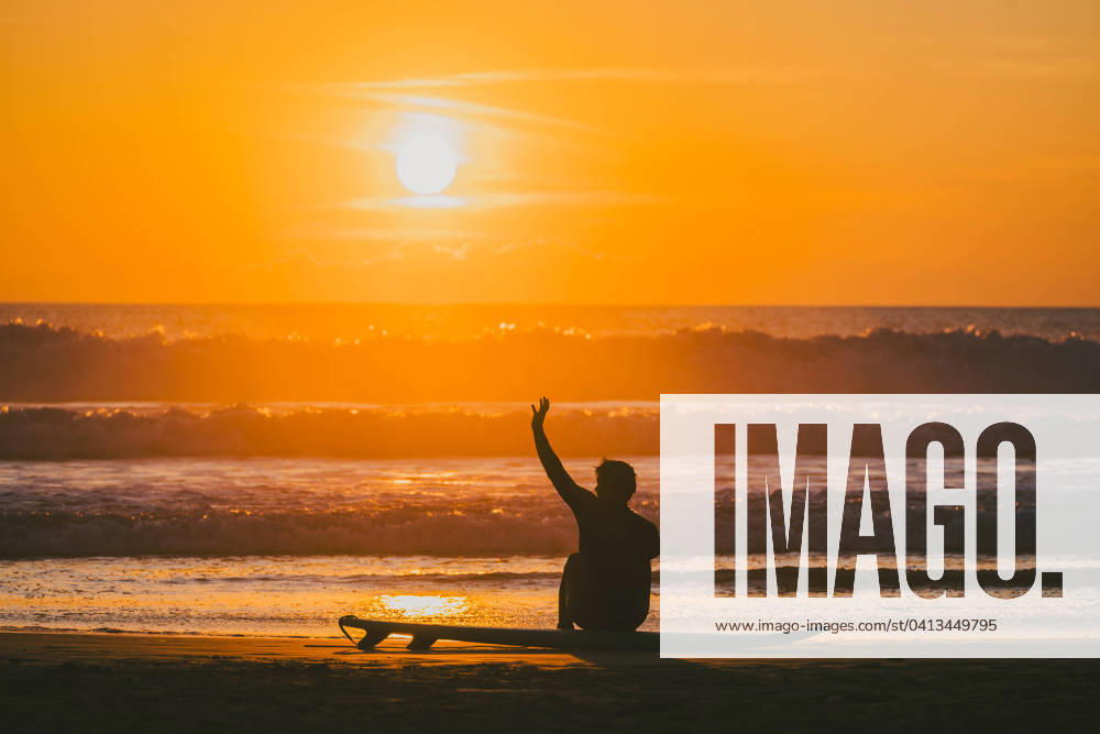 Unrecognizable male surfer sitting with raised hand on surfboard by sea at sunset Silhouette of unrecognizable male surfer sitting on surfboard with raised arm while enjoying picturesque sunset view over waving sea at evening Model Released AlvaroMontero_ID19256_573102_004