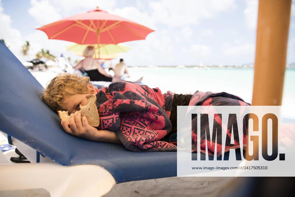 Boy Holding Sandwich sits on Lounge Chair Under Beach Umbrella Blowing Point Village, Anguilla CR_EMJA240225-109049-01 ,model released, Symbolfoto