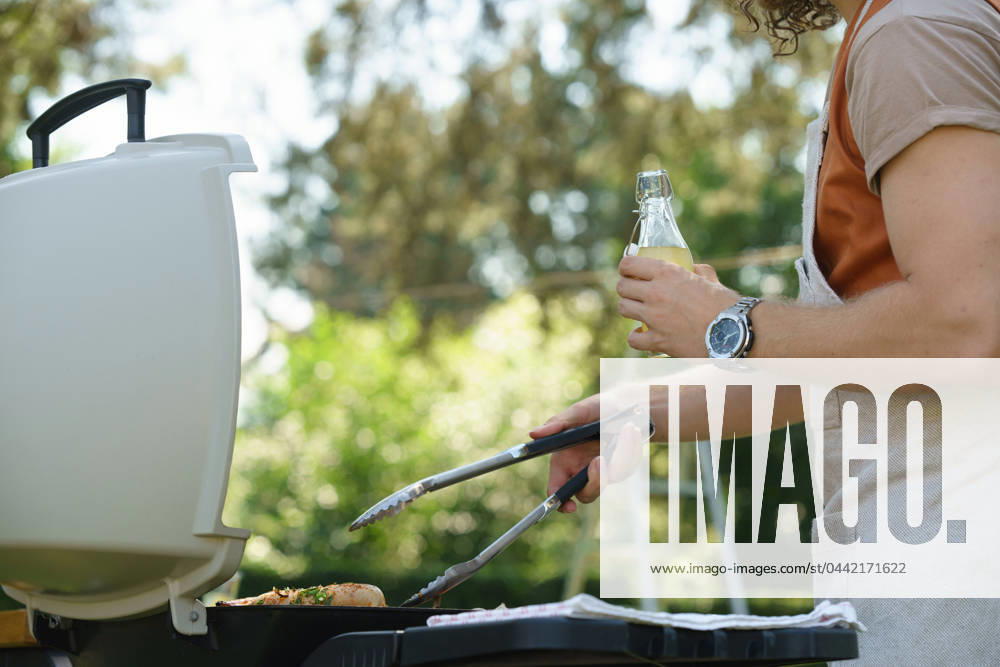 Man grilling on outdoor grill and drinkig lemonade. Outdoor grill or BBQ party in the garden. Man grilling on outdoor grill, holding tongs and drinkig lemonade. Outdoor grill or BBQ party in the garden. model released