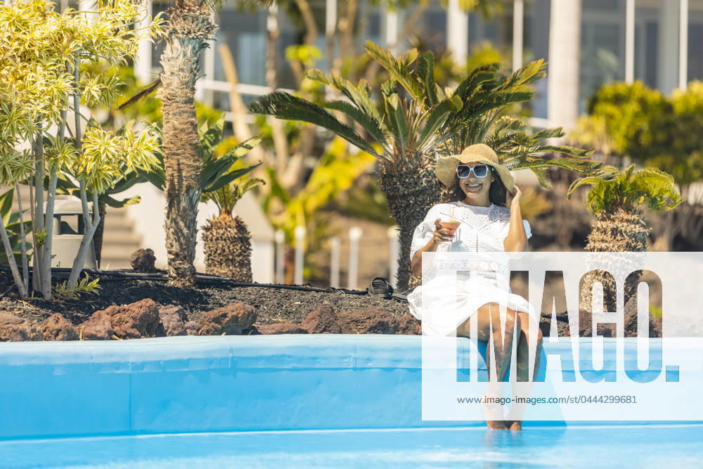 A smiling middle aged hispanic woman in a white dress and sunhat sits at the edge of a pool, holding a cocktail, surrounded by exotic plants. Model Released JuanAizpuru_ID20788_613331_028