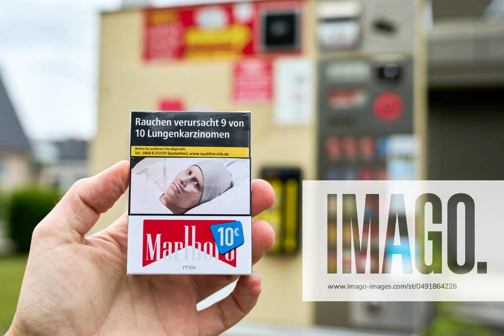 Hand hält eine Zigarettenschachtel von Marlboro zum Preis von 10 Euro vor einen Zigarettenautomat. Auf den Zigaretten ist ein Warnhinweis: Rauchen verursacht 9 von 10 Lungenkarzinomen