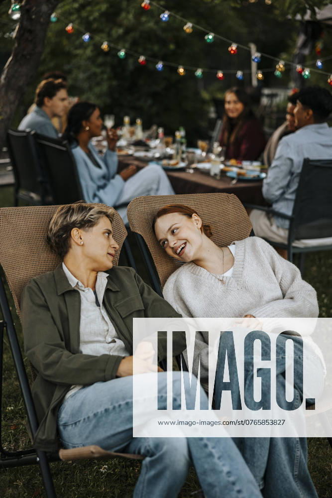 IMAGO Creative, Lifestyle, Feste im Garten #2 Female friends with legs crossed at knee sitting on chair and talking in back yard at social party Sweden, Stockholm, model released, property released
