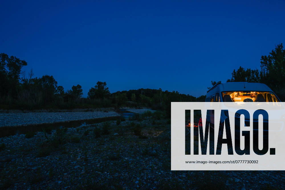A man sits on a camping chair in front of a table on a blue hour summer evening in front of his campervan Germany On the road in a campervan