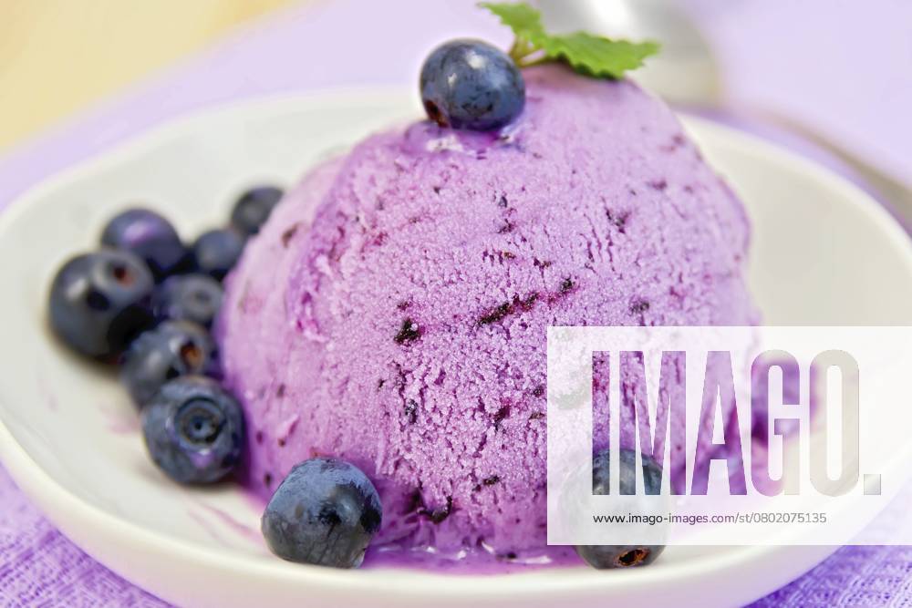 Heidelbeereis mit Minzblättern in einer Schale mit Beeren auf einem Hintergrund aus lila Stoff und Holzbrettern Blueberry ice cream with mint leaves in a bowl with berries on a background of purple cloth and wooden boards