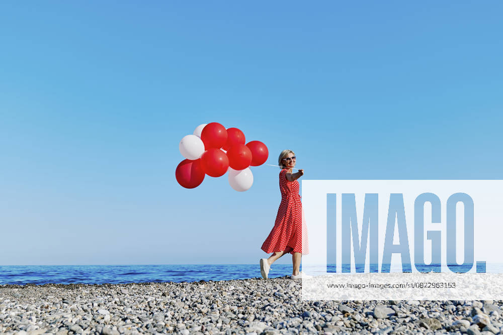 Blond woman in red polka dot dress holding balloons on a pebble beach by the Ionian Sea on a sunny day. model released, Symbolfoto, CATF00697