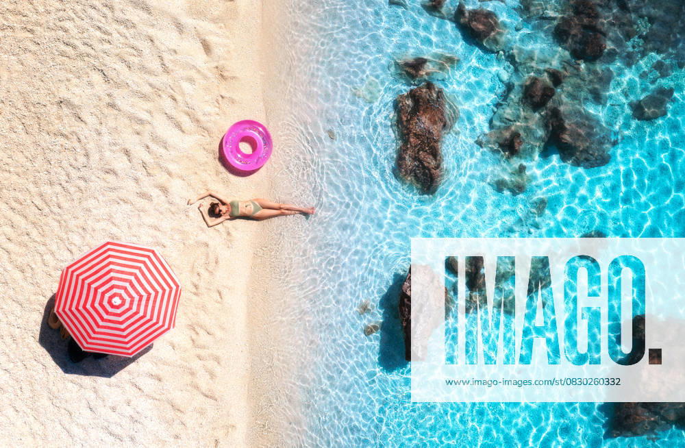 Aerial view of the lying beautiful woman with pink swim ring on the sandy beach, blue sea, stones, red umbrella at sunset. Summer vacation. Lefkada island, Greece. Top view of slim girl, azure water Model Released DenisBilytskyi_ID30691_1038775_024