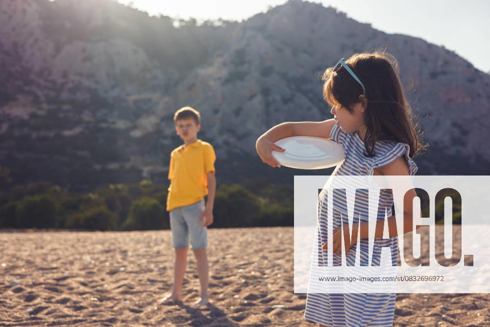 Children playing with frisbee on a sunny beach day A girl in a striped dress and a boy in a yellow shirt enjoy playing with a frisbee on a sandy beach. The sunny backdrop features rugged mountains and blue skies Model Released AlinaKandybka_ID31083_1054465_003