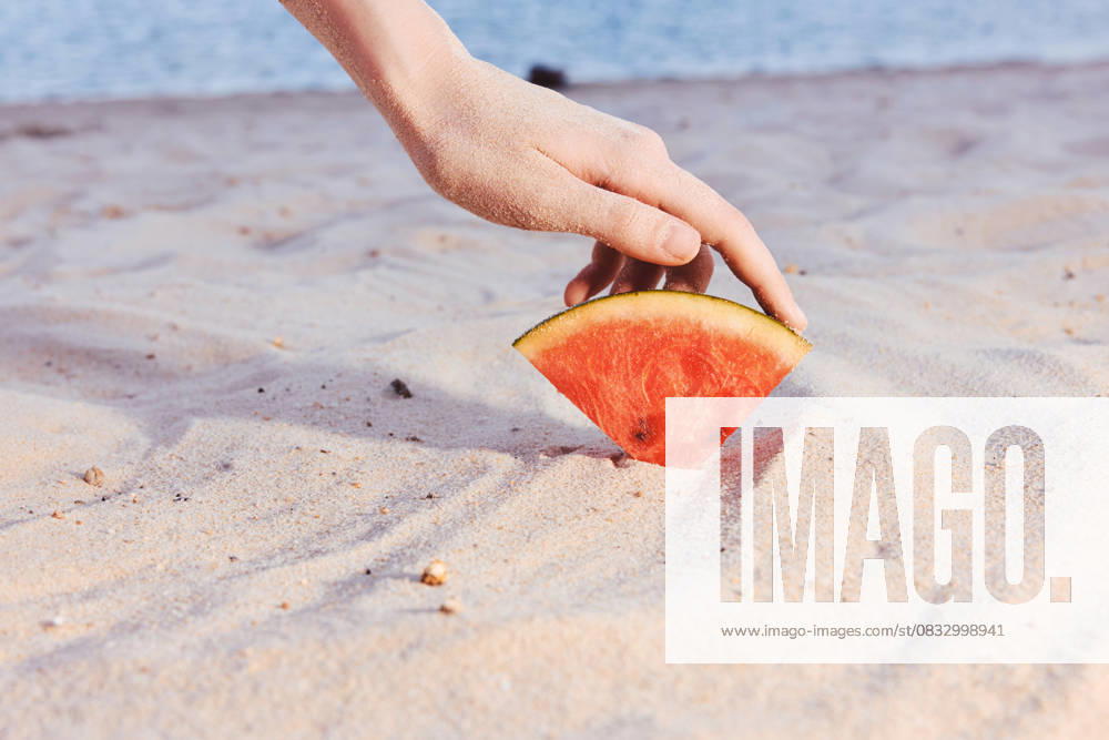 A hand placing a slice of watermelon on a sandy beach background, evoking summer and refreshment by the sea A hand gently places a slice of watermelon onto light colored sand at a beach, symbolizing summer, relaxation, and freshness, model released, , 96058526.jpg RECORD DATE NOT STATED