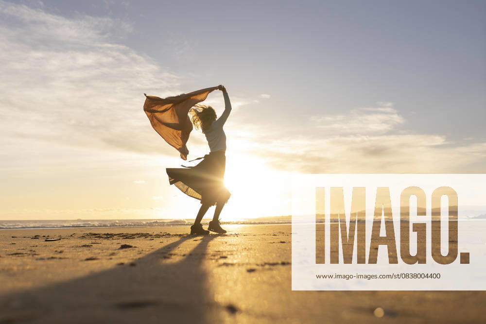 A woman stands on a beach with a scarf around her neck A woman stands on a beach with a scarf around her neck. The sky is a beautiful orange color, and the sun is setting. The woman is enjoying the moment and the beautiful scenery, 60897446.jpg RECORD DATE NOT STATED