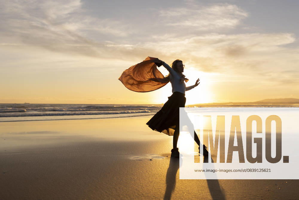 A woman is dancing on the beach with a scarf in her hand A woman is dancing on the beach with a scarf in her hand. The sky is orange and the sun is setting, model released, , 60777262.jpg RECORD DATE NOT STATED
