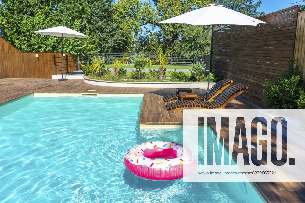 Hinterhofpool mit türkisfarbenem Wasser, Holzdeck, Sonnenschirmen, Liegestühlen und rosafarbenem Donut Float Backyard swimming pool with turquoise water, wooden deck, umbrellas, lounge chairs, and pink donut float