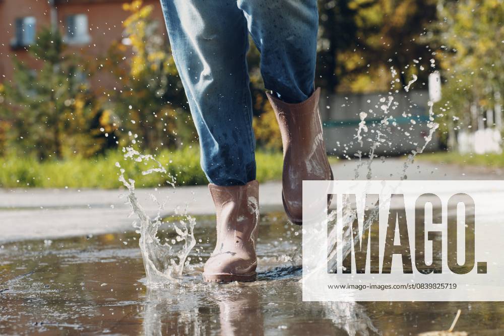 Frau trägt Regengummistiefel, läuft und springt in eine Pfütze mit Wasserspritzer und Tropfen im Herbstregen Woman wearing rain rubber boots walking running and jumping into puddle with water splash and drops in autumn rain