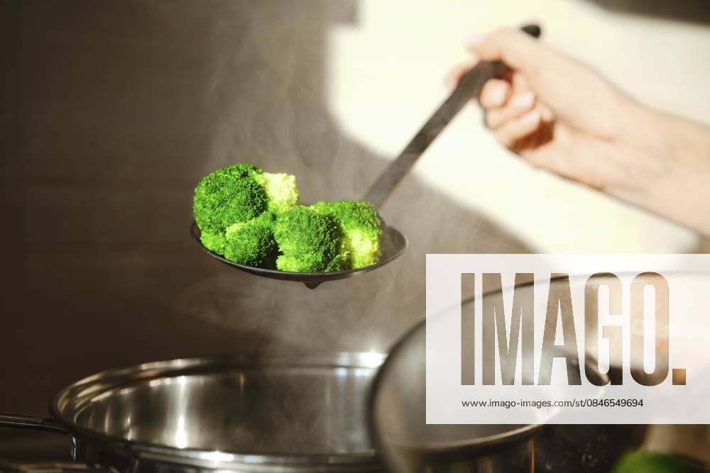 Eine Person hält einen Schaumlöffel mit gedämpftem Brokkoli über einen Topf in einer Küche. Der Dampf steigt auf und zeigt die Hitze, die beim Kochen entsteht A person holds a slotted spoon with steamed broccoli above a pot in a kitchen. The steam rises, showing the heat from cooking