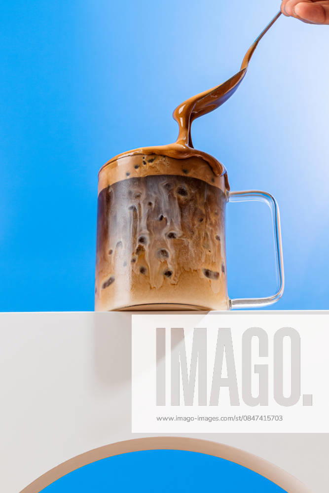 IMAGO Creative, Food, Trend, Iced Coffee Milky coffee action shot with spoon and glass mug An action shot capturing a creamy affogato pouring into a clear glass mug, with swirls of milk blending into rich coffee against a vibrant blue background. Model Released CintiaViudez_ID32864_1119261_002
