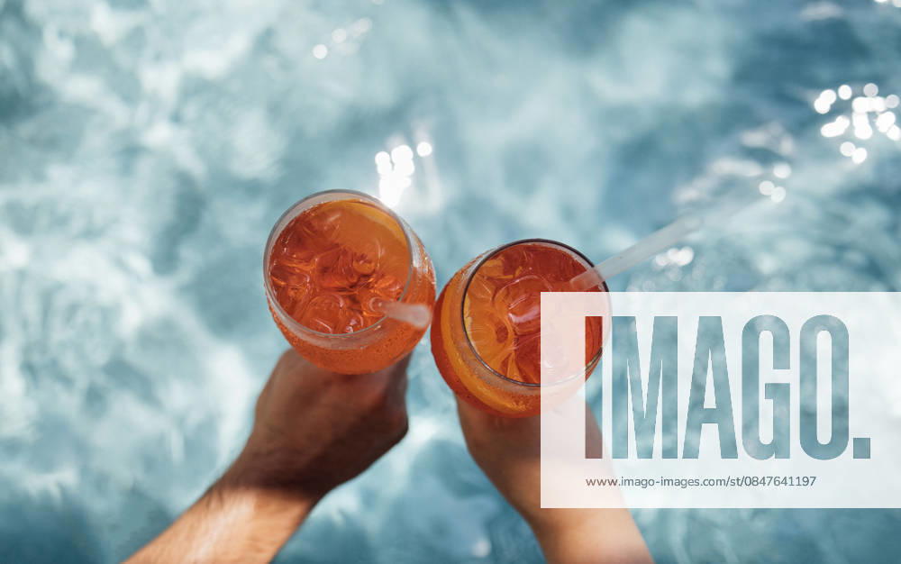 Zwei Hände halten Gläser mit orangefarbenen Cocktails mit Eiswürfeln, die teilweise in einem klaren Swimmingpool eingetaucht sind, Sonnenlicht reflektiert auf der Wasseroberfläche. Two hands grasp glasses of aperol spritz filled with orange cocktails and ice, partially submerged in a clear pool, with sunlight creating reflections on the water (License=RF)