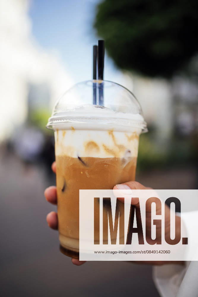 IMAGO Creative, Food, Trend, Iced Coffee Eiskaffee in einem durchsichtigen Plastikbecher, gehalten von einer Hand mit zwei schwarzen Strohhalmen, verschwommener städtischer Hintergrund mit Grünflächen und vorbeigehenden Menschen Iced coffee drink in a clear cup with whipped cream and two straws, held by a hand against a blurred urban setting with greenery and pedestrians (License=RF)