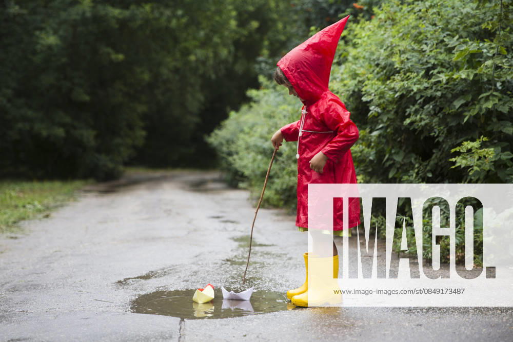 Ein Kind in einem roten Regenmantel und gelben Gummistiefeln spielt nach einem Sommerregen mit Papierbooten in einer Pfütze. a child in red raincoat and yellow rubber boots playing with paper boats in a puddle after rain in the summer (License=RF) ,model released, Symbolfoto