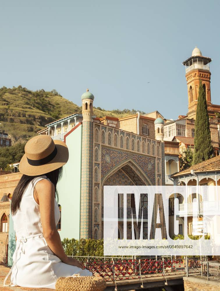 Frau sitzt im Sommerkleid mit Strohhut und bewundert die lebendige Architektur der Altstadt von Tiflis, Georgien. Traditionelle Holzbalkone, Moschee Minarett und historische Gebäude. Reisen, Sightseeing, Sommerurlaub Woman sit in summer dress straw hat admiring vibrant architecture old town Tbilisi, Georgia. Traditional wooden balconies, mosque minaret, and historic buildings. Travel, sightseeing, summer vacation