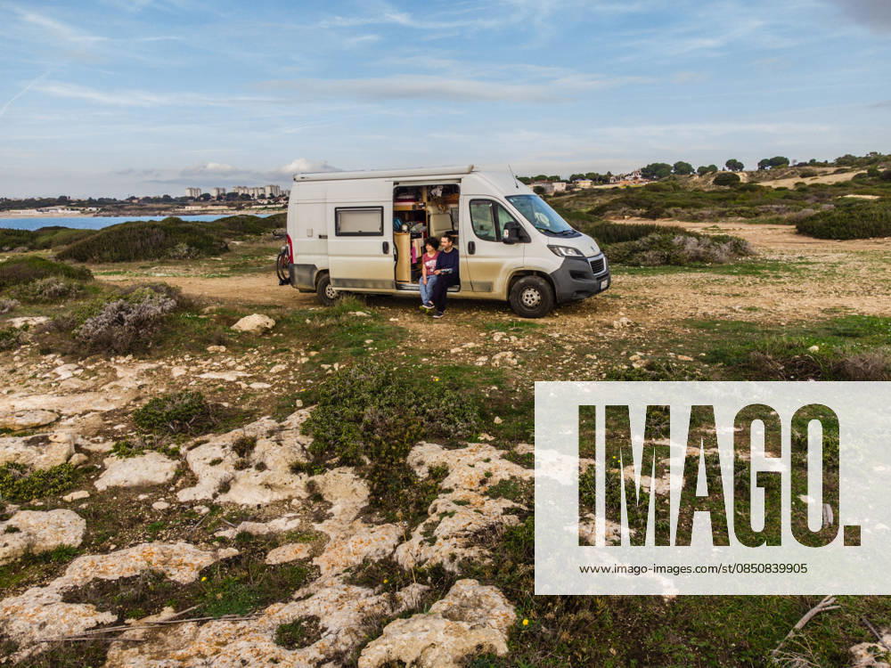 Campers sit in the sliding door of their camper on an unpaved off-road pitch on the Mediterranean coast near Taranto Salento, Puglia, Italy A camper van is parked on a gravel pitch on the shore of the Mediterranean Sea