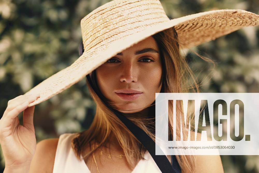 Porträt einer jungen stilvollen Frau, die ihren Strohhut hält, während sie vor einer grünen Wand steht Portrait of young stylish woman holding her straw hat while standing in front of a green wall