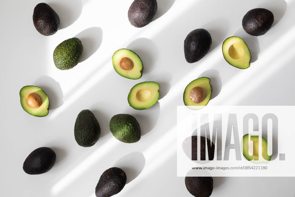 Ein Haufen reifer ganzer und halber Avocados auf weißem Hintergrund, von oben betrachtet, in ästhetischem Schattenlicht. Flat-Lay-Stil. Heap of ripe whole and half avocados on white background top view in aesthetic shadow sunlight. Flat lay style. (License=RF)