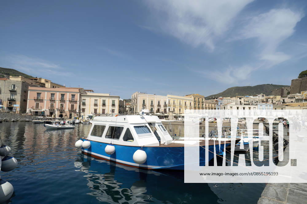 Ein traditionelles blau-weißes Fischerboot, das im Hafen von Mondello auf Sizilien vor Anker liegt. Bunte historische Gebäude säumen die Uferpromenade am Mittelmeer. Eine Szene aus einem italienischen Küstendorf. Traditional blue and white fishing boat moored in Mondello harbor, Sicily. Colorful historic buildings line the waterfront with Mediterranean Sea. Italian coastal village scene. (License=RF)