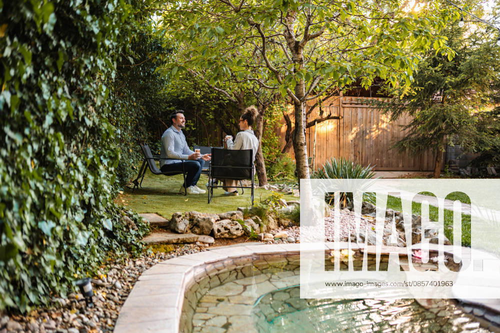 Ein Paar, das sich im Garten hinter dem Haus entspannt und einen Kaffee genießt Couple sitting at a table with drinks in a lush backyard garden with a pond (License=RF) ,model released, Symbolfoto