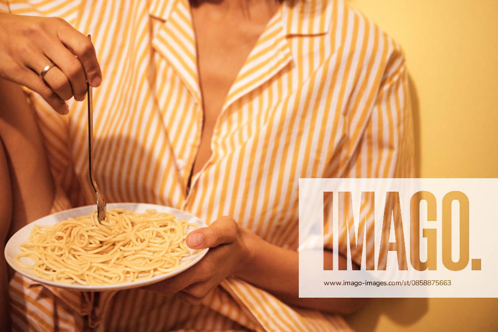 Eine Frau in einem gestreiften Pyjama sitzt an einer gelben Wand und hält einen Teller Spaghetti mit einer Gabel in der Hand; die Szene vermittelt den Eindruck eines ungezwungenen Essens zu Hause Woman in striped sleepwear enjoys a plate of spaghetti while seated against a yellow wall, creating a cozy home dining atmosphere (License=RF) ,model released, Symbolfoto