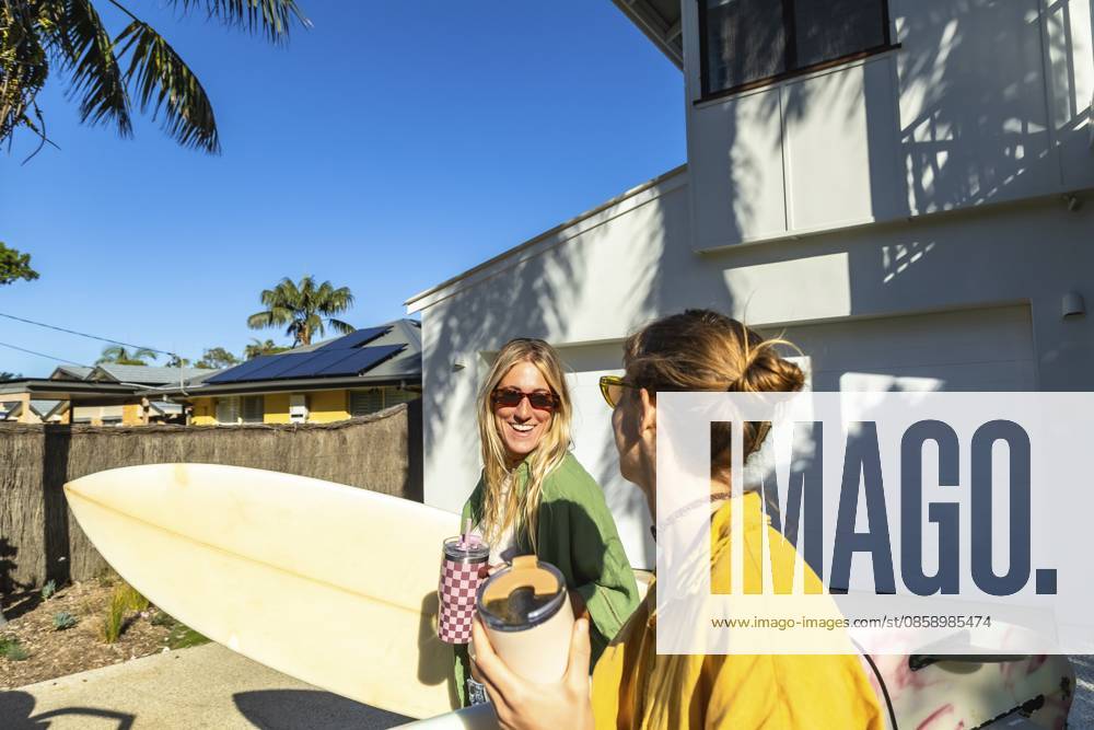 Zwei junge australische Surferinnen stehen draußen mit Surfbrettern, halten Getränke in der Hand und unterhalten sich unter dem strahlend sonnigen Himmel, um die Küstenkultur und den Sommergenuss zu genießen Two young australian women surfers standing outdoors with surfboards, holding beverages, and engaged in conversation under the bright sunny sky, embracing coastal culture and summer enjoyment